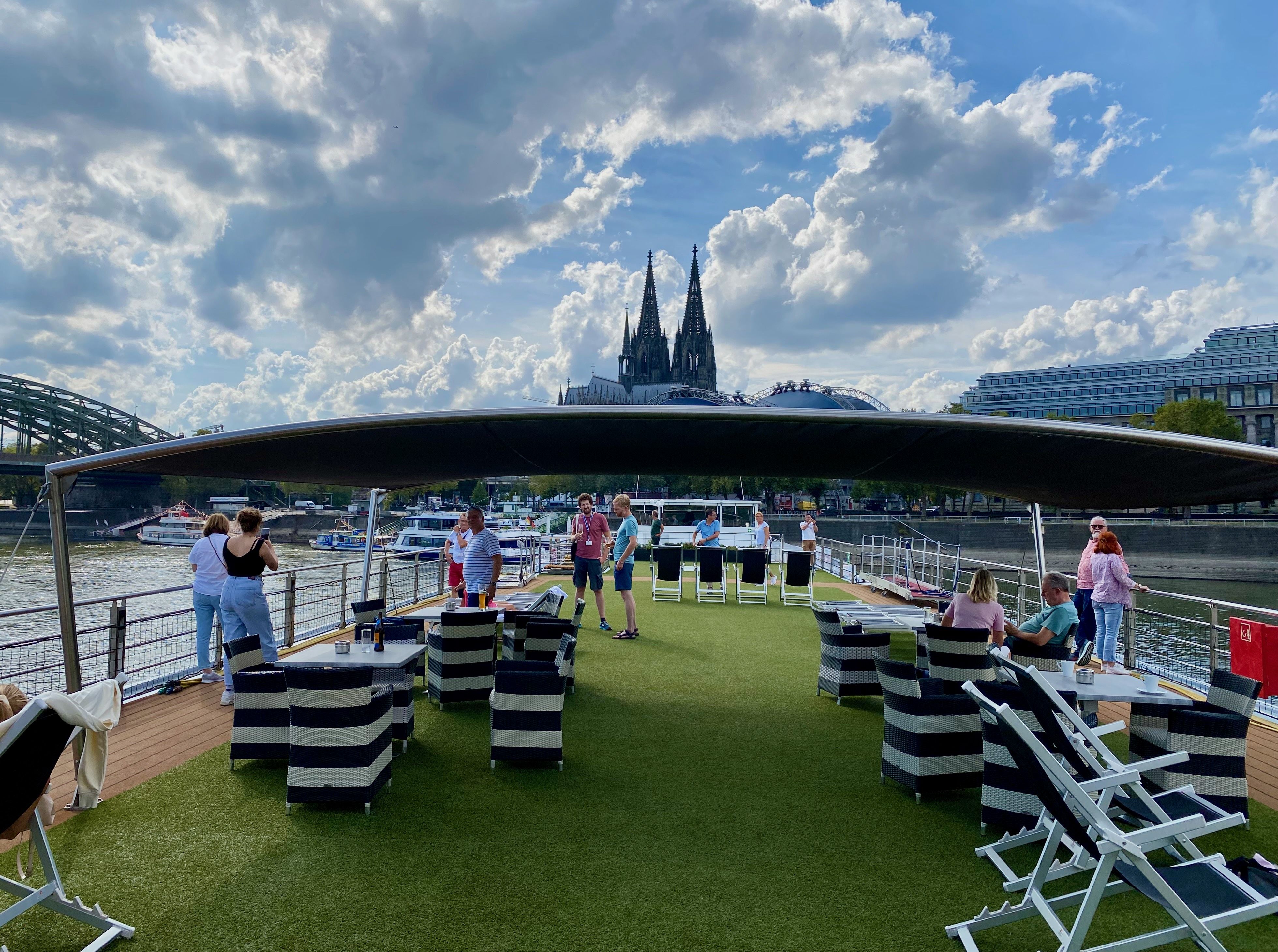 Sonnendeck eines Flusskreuzfahrtschiffs mit Sitzbereichen und Gästen vor dem Kölner Dom während einer Reise mit Hund