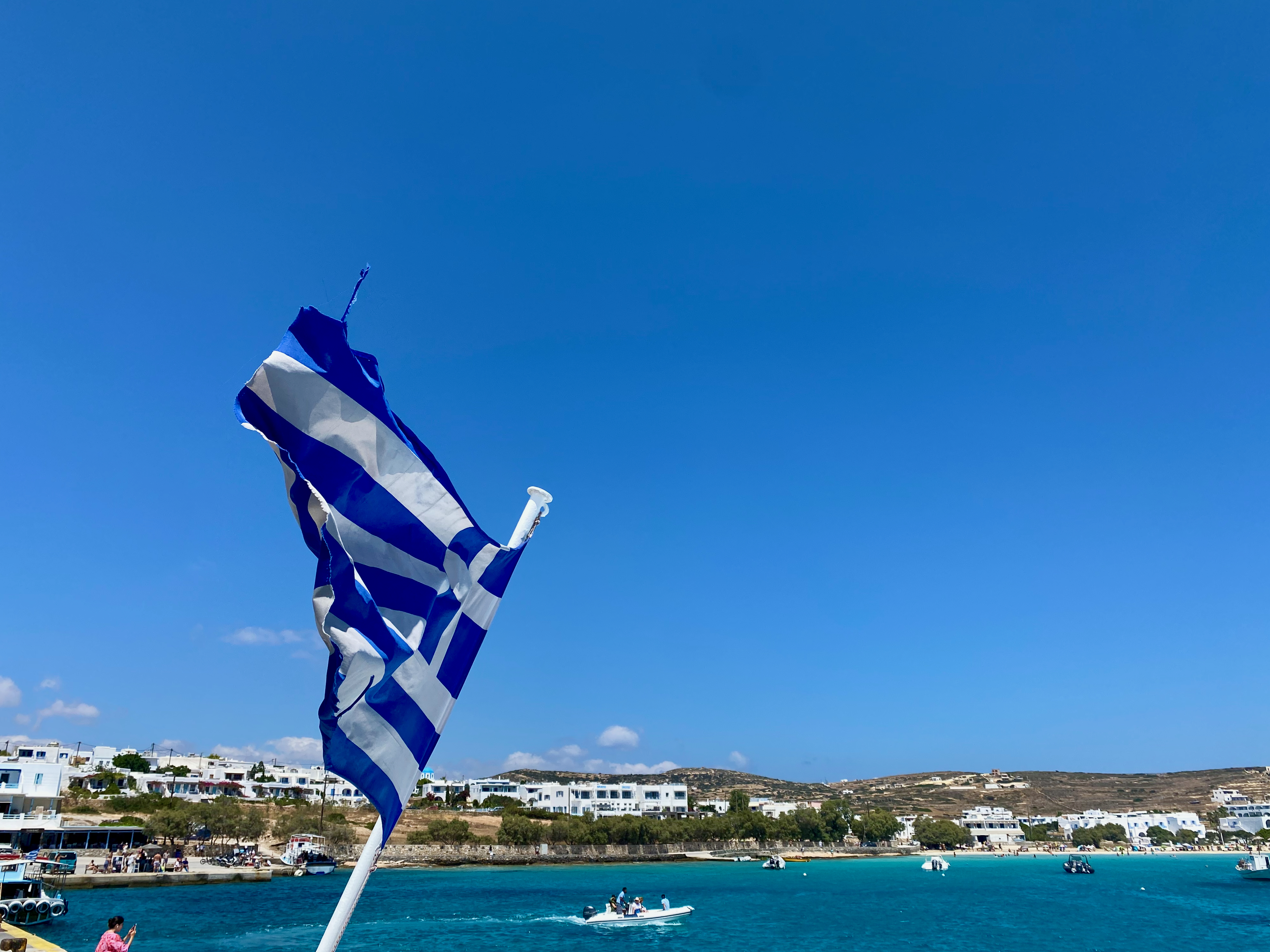 Griechische Flagge weht im Wind vor einem Hafen in den Kykladen mit Meer und Küstenort